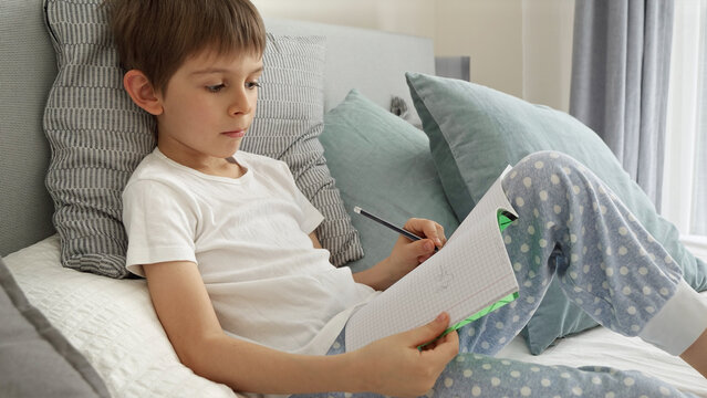 Portrai of little boyworking on his homework in bed, writing in a notebook with concentration. Importance of education and child development in a remote learning environment.