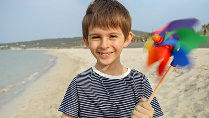 Portrait of cheerful smiling boy having fun on sea beach with colorful pinwheel. Concept of summer holiday, happiness, joy, travel and weekend.