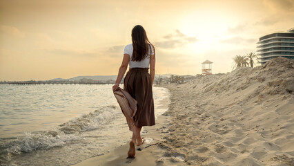 Woman walking on the sandy sea beach at a windy day, holding a silk scarf. Beauty and peacefulness of a summer holiday.
