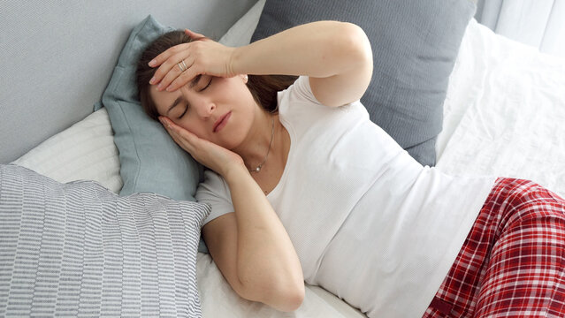 Young sick woman having high temperature lying in bed and holding hand on forehead while suffering from headache