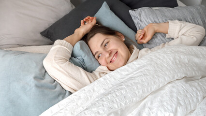 Portrait of smiling young woman opens eyes while lying and waking up in bed at morning. Concept of comfort, relaxation, healthy sleeping and good start of new day.