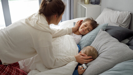 Obraz premium Portrait of smiling loving mother covering with blanket and looking at her children sleeping in bed. Concept of family happiness, relaxing at home, having fun in bed, parent and cheerful kids