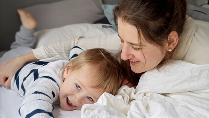 Young loving mother embracing her cute toddler son lying in bed at morning. Concept of family happiness, relaxing at home, having fun in bed, parent and cheerful kids