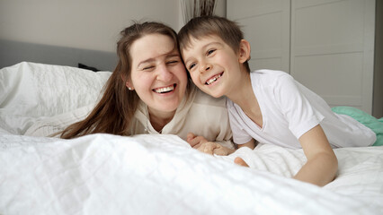 Obraz na płótnie Canvas Cheerful boy with mother in pajamas lying on soft bed at bedroom. Concept of family happiness, relaxing at home, having fun in bed, parent and cheerful kids
