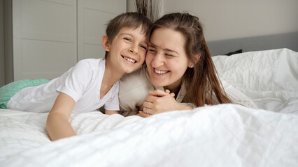 Obraz na płótnie Canvas Happy cheerful boy with mother wearing pajamas lying in bed and having good time together