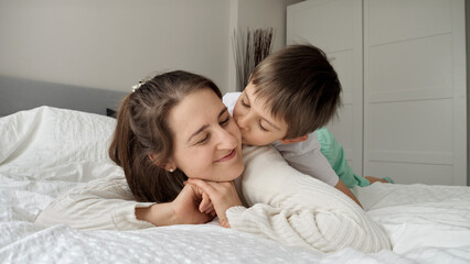 Obraz na płótnie Canvas Cute loving boy in pajamas lying with mother in bed and kissing her. Concept of family happiness, relaxing at home, having fun in bed, parent and cheerful kids