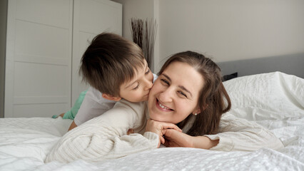 Portrait of cute boy lying in bed with mother and kissing her in cheek