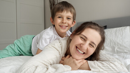 Happy cheerful boy lying with mother in bed and hugging her. Concept of happy family, parents with kids, positive emotions and relaxing at home.