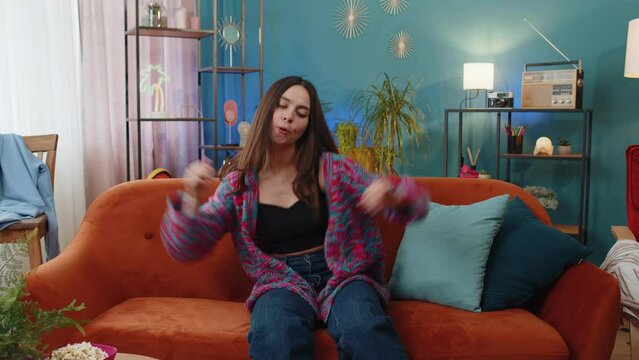 Trendy cheerful positive young woman having fun dancing and moving to rhythm, dabbing raising hands, making dub dance gesture. Portrait of Caucasian girl celebrate win at home apartment room on sofa