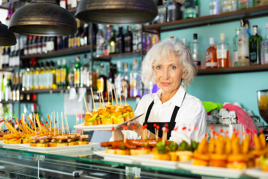 Positive mature female barman in uniform serving delicious pinchos at the counter of the bar. High quality photo