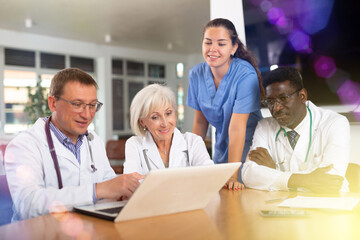 Obraz premium In morning meeting, doctors of emergency department confer about patients clinical case. Male health worker with colleagues sitting at table, reading new article on laptop and discussing information