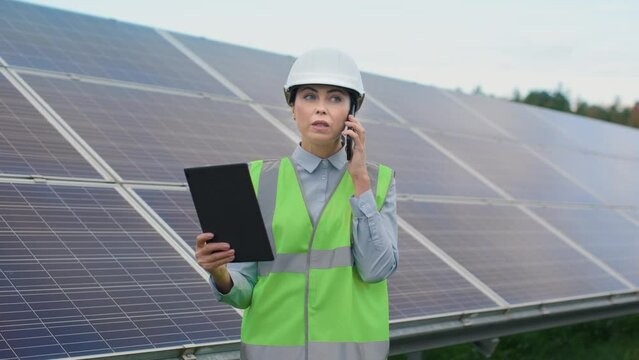 Portrait of female worker in spesial uniform and hard hat on background of solar panels. Ecological engineer is takling by phone and works with using of tablet outside. Green electricity concept.