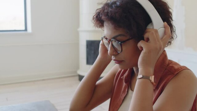 Close-up tilting shot of young African American woman with curly hair, in glasses and sleeveless shirt sitting at table at home, typing on laptop, then adjusting large white headphones