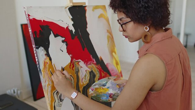 Medium arc shot of young multiracial woman with dark skin and curly hair standing in front of easel at home, holding colour palette and putting finishing touches to bright abstract painting
