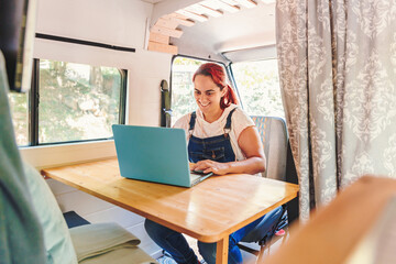 Caucasian young woman working on her laptop in her handmade camper van while traveling. Van road...