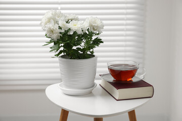 Beautiful chrysanthemum plant in flower pot, cup of tea and book on white table indoors