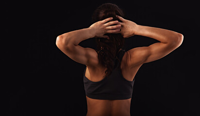 Female sporty muscular with ponytail doing stretching workout of the shoulders, blades in sport bra, holding the neck hands on black background with empty copy space. Back view.