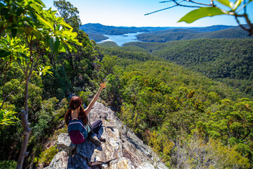 Naklejka na ściany i meble Beautiful girl sits on a rock shelf enjoying view of the Advancetown lake after successfully climbing the Pages Pinnacle. Hidden gems in Springbrook National Park, Gold Coast, Queensland, Australia