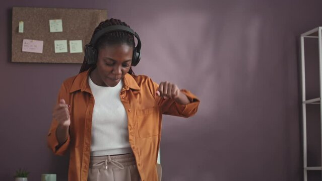 Medium shot of young vibrant African American woman dancing to music in headphones while working with brand marketing from home