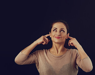 Beautiful business happy smiling woman covering the ears the fingers to not want listening sounds and dreaming looking up on studio black background. Closeup