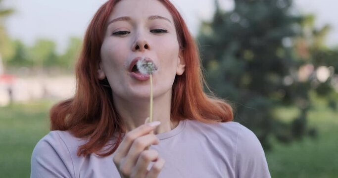 The joyous moments of a young woman blowing on a dandelion she found in the park and directing it towards the camera capture a sense of whimsy and hope