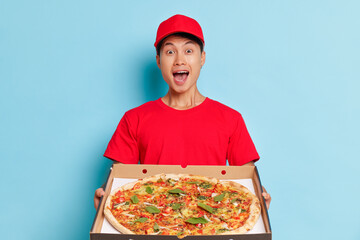 Happy cute young man in red clothes and cap stands on blue background with box of large pizza in...