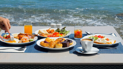 Delicious buffet breakfast served with a variety of eggs, sausage, coffee, fresh orange juice, croissants, and salad with sea view. Man enjoys his meal, dining outside, enjoying idyllic ocean view.