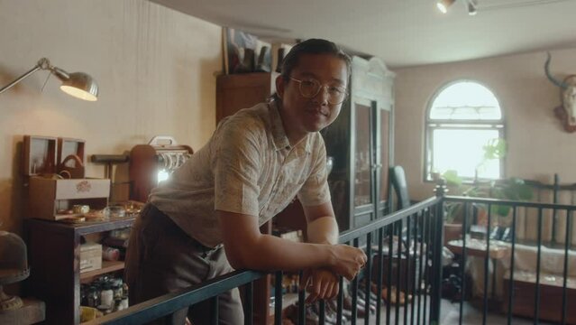Young Asian antiques collector standing in vintage store, leaning on railings, posing for the camera with smile