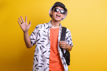 Young cheerful Asian backpacker wear casual clothes, sunglasses and bucket hat waving hand, welcoming and greeting you, or saying goodbye against yellow wall