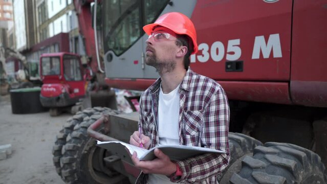 Young construction manager checks the technical documentation while on construction site near heavy construction equipment tractor or excavator. Foreman checking report.