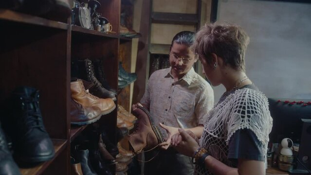 Asian salesman telling about old leather boots to young female customer, helping her with choice in vintage store of secondhand and rare items