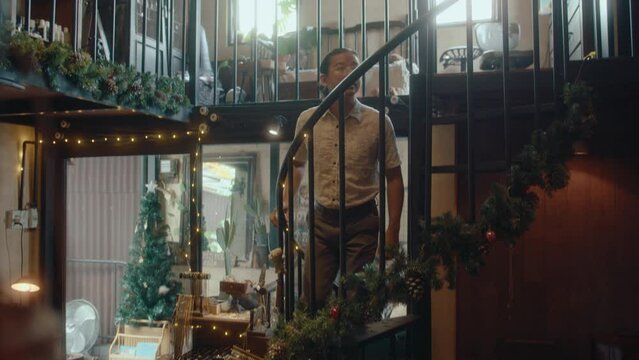Asian man walking up on spiral staircase to the second floor of antique shop decorated with lights and fir branches on Christmas