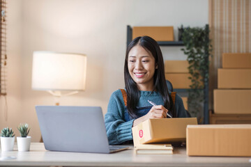 Young beautiful happy asian business woman owner of SME online using laptop receive order from customer with parcel box packaging at her startup home office, online business seller