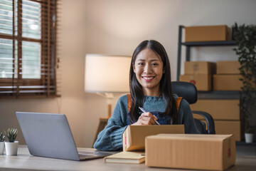 Naklejka premium Young beautiful happy asian business woman owner of SME online using laptop receive order from customer with parcel box packaging at her startup home office, online business seller