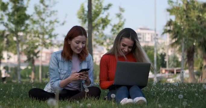 The two young women, sitting in the park, engrossed in their laptops and phones, diligently working on their tasks, become captivated by the scenery and derive immense pleasure from it