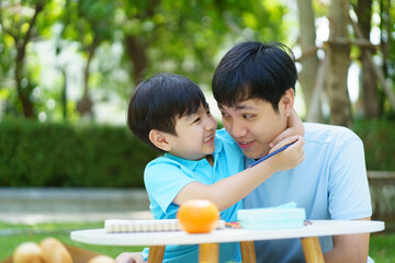 Happy lovely Asian father and son enjoy relaxing in picnic in the garden together, boy hugs his daddy. Dad and son having a meal and picnic in garden.