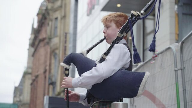 Young man wearing tartan is playing bag pipes outside large buildings