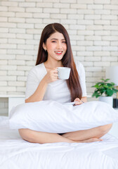 Portrait closeup shot Millennial Asian beautiful cheerful happy female model in casual outfit sitting smiling posing holding hot coffee cup and dish on white pillow look at camera on bed in bedroom