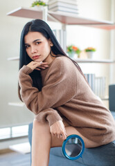 Millennial Asian beautiful pretty female model in casual outfit big brown warm sweater sitting on cozy sofa couch holding handheld mirror posing in living room.