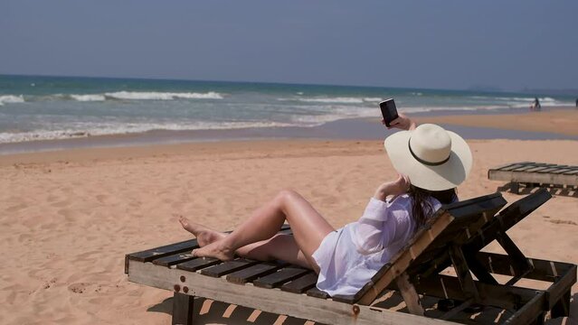 Woman tourist in sun hat making selfie on smartphone, back view. Brunette female lying sitting sunbed on sandy ocean beach resting on tropical resort. Tourism travel, vacation, photographing moments.