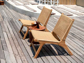 Two empty modern cozy brown rattan chairs decoration on outdoor wooden deck on summer sunny day. Twin wooden seats with side table with no body.