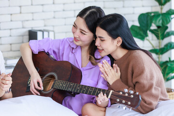 Closeup shot of Millennial two Asian beautiful cheerful female LGBTQ lover couple girlfriends in casual outfit sitting cuddling embracing smiling holding playing guitar singing songs music together