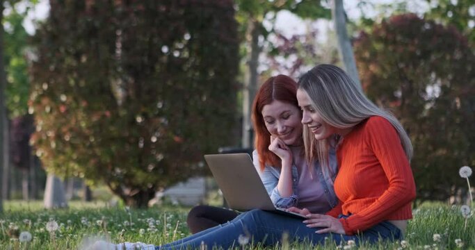 The two young women, sitting in the park, engrossed in their laptops and phones, diligently working on their tasks, become captivated by the scenery and derive immense pleasure from it