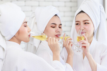 Three Asian cheerful happy beautiful female girlfriends in casual bathrobe and towel outfit standing smiling holding sparkling champagne tall glass cheers toasting together in spa massage studio