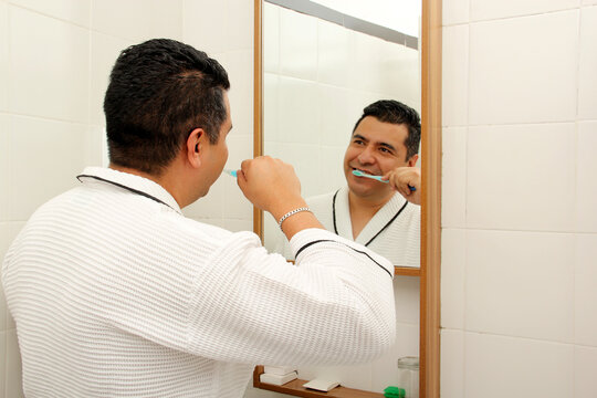 Forty-year-old dark-skinned Latino man brushes his teeth in the bathroom in a bathrobe looks in the mirror and takes care of his oral health
