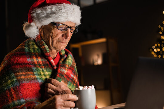 Side view of old man drinking cocoa with marshmallows on christmas eve, Elderly man in santa hat alone watching movie on internet at home