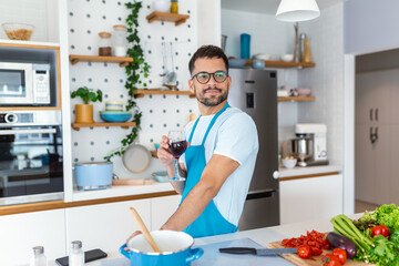 Home cook. Single man in apron looking at the window, drinking wine from a glass while cooking in the kitchen, cooking at home