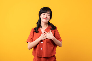 Portrait young beautiful asian woman happy smile dressed in orange clothes showing hands touching her chest in proud and thankful emotional feeling isolated on yellow studio background.