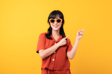 Portrait young beautiful asian woman happy smile dressed in orange clothes and sunglasses showing pointing finger hand gesture to free space isolated on yellow studio background. Attention to product