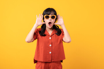 Portrait young beautiful asian woman happy smile dressed in orange clothes and sunglasses looking surprised, reacting amazed, raising eyebrows impressed isolated on yellow studio background.